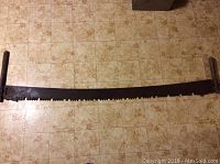 Full length of the antique two-person crosscut saw laid on a beige patterned tiled floor.