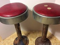 Front and side view of two vintage stools with red leather tops and round metal bases, showing rust and wear.