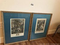 Photo of two large framed vintage engraving prints leaning on the wall showing detailed artwork and blue matting under glass with ornate gold colored frames.