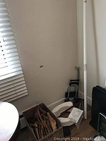 Corner with white toilet seat on floor, wooden hangers in box, and vertical white safety post against wall