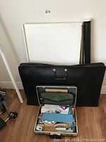 Photo showing a large black carrying case, drafting table top, rolled paper tube, and a metal briefcase with various drafting supplies inside.