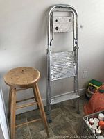 Small aluminum step ladder and wooden stool positioned against a wall on a tiled floor