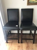 Two black leather stools standing side-by-side showing their front and side profiles, placed on a wooden floor near a window.