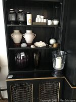 Shelf containing two white urn-style vases, dark glass vases, large clear glass candle holder with candle, shells, and tea candles on upper shelf.