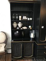 Front view of black and gold painted pine cupboard with brass grilled doors, shelves populated with jars and decorative items.