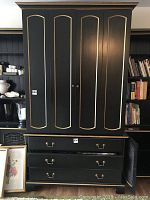 Front view of entertainment unit showing four tall paneled doors with brass knobs and three drawers below with brass handles. Black with gold trim.