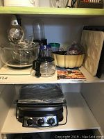 Wide angle view of Sunbeam stand mixer with glass mixing bowls, Magic Bullet blender with pitcher and cups, and panini grill with plastic wrapped top.