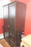 Full view of tall dark faux wood cabinet showing two doors and two bottom drawers with metal handles. Positioned against a red wall.