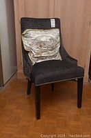 Front angled view of dark gray upholstered chair with gold decorative pillow on seat and visible nailhead trim on the bottom edge