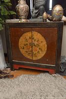 Front view of wooden cabinet showing circular floral painting on front doors with two metal pulls and base painted red.