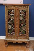 Front view of carved wood cabinet with detailed floral and leaf carvings, green and reflective decorative accents, standing on four carved legs with scalloped base.