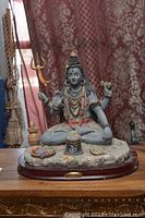 Front view of Lord Shiva figurine showing seated posture with trident and instruments, set against a patterned curtain background.