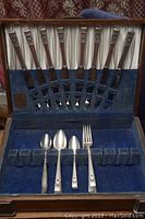 Full view of vintage flatware set in blue lined case showing 8 knives arranged in top row and some spoons and a fork in bottom row, with noticeable wear to case lining and slots.