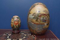 Two Asian style decorative eggs on a patterned surface against a blue wall, larger egg standing and smaller egg on a wooden stand
