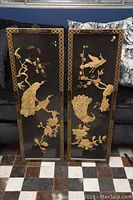 Two lacquered wood panels side by side on floor against sofa, showing overall design of carved birds and floral elements on black lacquer background with patterned border.