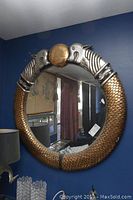 Full view of round mirror showing frame with fish heads and scaled body against blue wall, reflecting room interior.