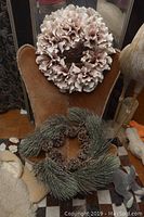 Two decorative wreaths placed on a chair; one consists of pinkish-beige fabric petals and the other has green artificial pine boughs with pine cones.