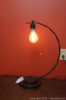 Full view of the black curved arm desk lamp lit up against a red wall, showing height and structure.