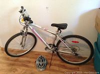 Full side view of the ladies Infinity Back Road silver and pink bike with front suspension fork, resting on kickstand, with Bell helmet placed on floor in front.