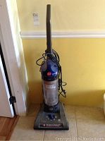 Full view of Hoover upright vacuum showing handle, power cord, and dust container placed against a wall on tiled floor.