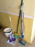 Photo showing Bissell Swiffer floor cleaner, broom, dustpan, a white bucket, and a pack of Swiffer refill pads against a yellow wall.