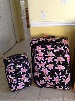 Two piece set of Aviator brand luggage, one large and one small, both black with pink and white floral pattern, shown side by side on floor.