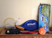 Photo of assorted outdoor toys on wood surface showing Wilson tennis racket in cover, Cooper baseball mitt, Ladder Ball game, badminton rackets with shuttlecock, and an orange ball.