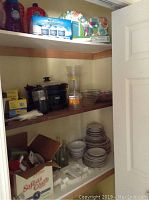 Shelves containing boxed food storage sets, black Coleman cooker, assorted bowls and plates stacked and organized, and a box with miscellaneous items including bottles and kitchen items.