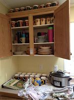 Cabinet with assorted mugs, plates, teapot and kitchen towels on countertop with cookbooks and Crockpot