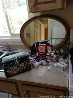 Photo showing large oval mirror with ornate embossed frame behind a countertop with jewellery and sewing kit box.