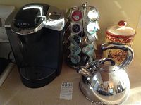 Photo showing Keurig coffee maker, Lagostina stainless steel tea kettle, ceramic cookie jar, and rotating coffee pod holder on countertop.