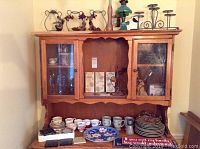 Wide view of kitchen hutch with decorative items on top and assorted tea cups, plates, mugs displayed on the bottom shelf along with plaques and small kitchenware items.
