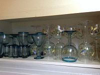 Shelf showing assortment of hand-painted wine glasses and blue margarita glasses with shot glasses.