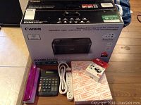 Canon Pixma MX492 printer box along with ink cartridge box, calculator, power cord, power strip adapter, and rewrapped ink paper shown on wooden floor.