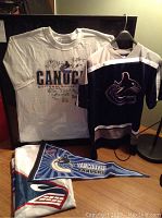Photo showing framed signed white Canucks tee-shirt, youth size 18 XL blue jersey with Orca logo, large flag, and pennant with Vancouver Canucks branding