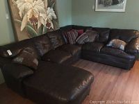 Wide angle view of large L-shaped dark brown leatherette sectional sofa with tufted back cushions and several decorative pillows in living room setting.