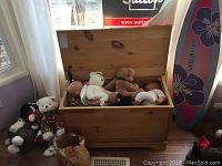 Photo showing wooden toy chest open with stuffed bears and baby dolls inside, along with some bears outside the chest, window and some floor visible