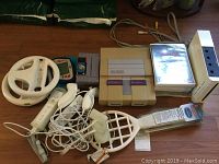 Photo showing the full lot including SNES console, Super Mario World cartridge, Wii console, two Wii controllers with Nunchuks, Wii Wheel, cables and game cases.