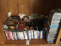 Photo of DVDs and CDs stacked in a wooden shelving unit showing multiple rows of DVDs in cases and some CDs on the left side.