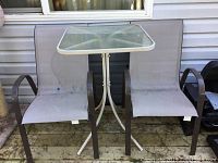 Table with glass top and two matching mesh sling chairs placed on a wooden deck near a windowed wall of a house.