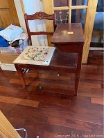 Photo of vintage wooden telephone table with attached chair showing embroidered floral seat and carved backrest. Visible marks, scratches, and wear on wood surface.