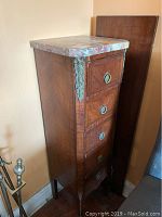 Full view of narrow antique wooden lingerie chest with marble top sitting near wall and other furniture.