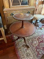 Full view of the two tier round wooden table with carved legs on a patterned rug, showing upper and lower tiers and overall shape.