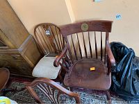 Photo showing two wooden chairs: one with cushioned seat and vertical slat back, one armchair with coat of arms plaque on backrest.