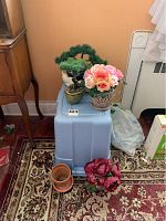Photo showing two faux bonsai trees, one faux floral arrangement in a decorative vase, a small terracotta clay pot, and a bag of potting soil arranged on top of and around a blue plastic container.