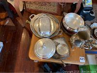Photo showing eight silver-plate pieces including pitcher, tray, covered dish, bowl, and small plates arranged on a wooden table.