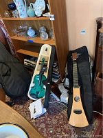 Wide view showing standing Martin Backpacker travel guitar in soft gig bag and turquoise violin in white hard case on carpet floor near wooden shelves with glassware.