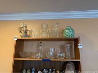 Shelving with various clear glass pitchers, crystal bowls, decanter, short glasses, and a green and white vase on the top two shelves. Small blue and white figurines on lower shelf.
