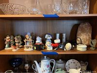 Shelf with mix of Hummel figurines, animal figures, glassware, and salt lamp.