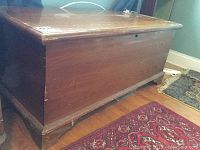 Front-side angle view of wooden cedar chest showing hinged lid and bracket feet on wood floor with nearby rug.